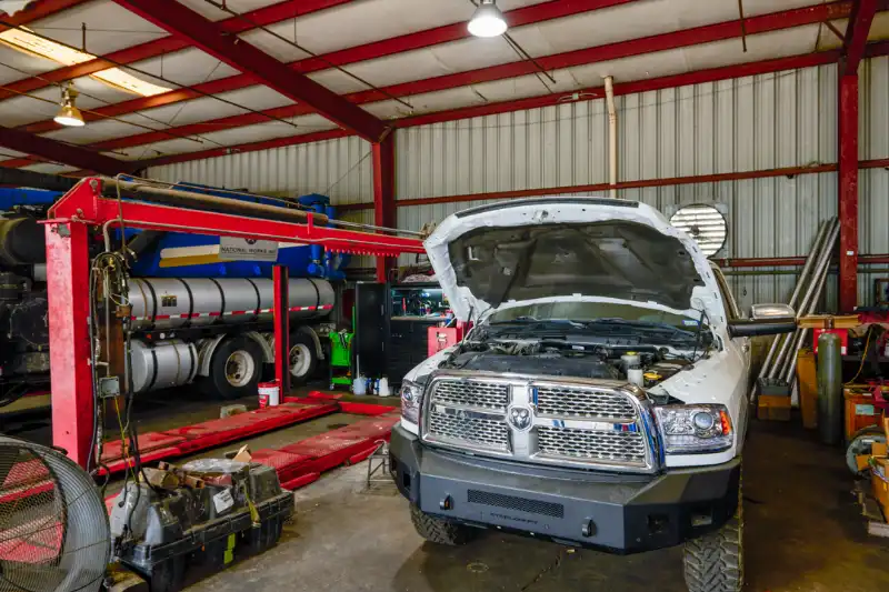 Mechanic in blue uniform working on a truck engine with the hood open inside a repair shop.